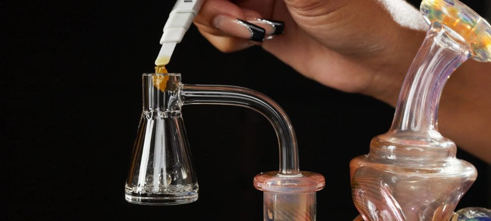 Gros plan d’une main plaçant un concentré sur un banger en verre transparent monté sur un dab rig, avec un fond sombre.